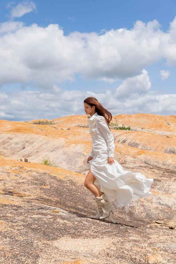 mulher em paisagem rochosa com vestido longo branco e bota bege de salto médio