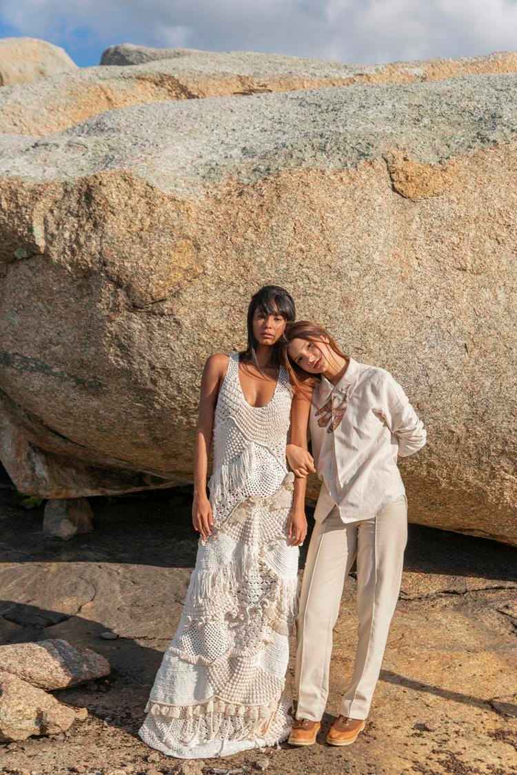 duas mulheres em paisagem rochosa, uma com vestido longo de renda branco e outra com camisa e calça de linho