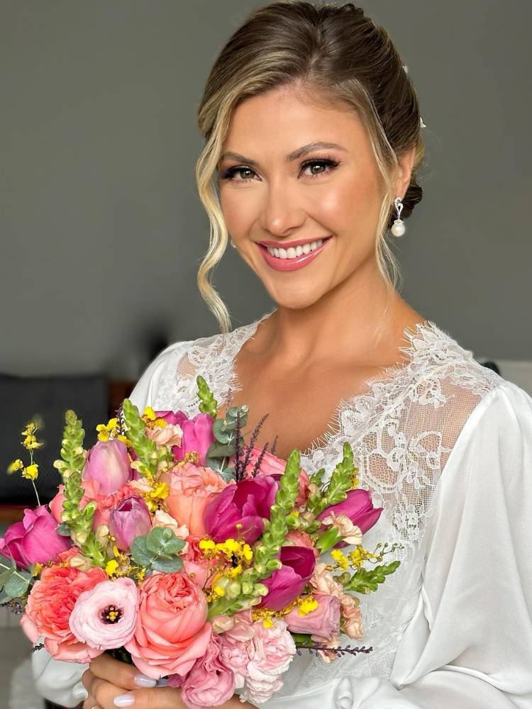 Noiva sorridente com make natural segurando buquê de flores variadas nas cores rosa, salmão e pink, usando vestido branco com detalhes de renda