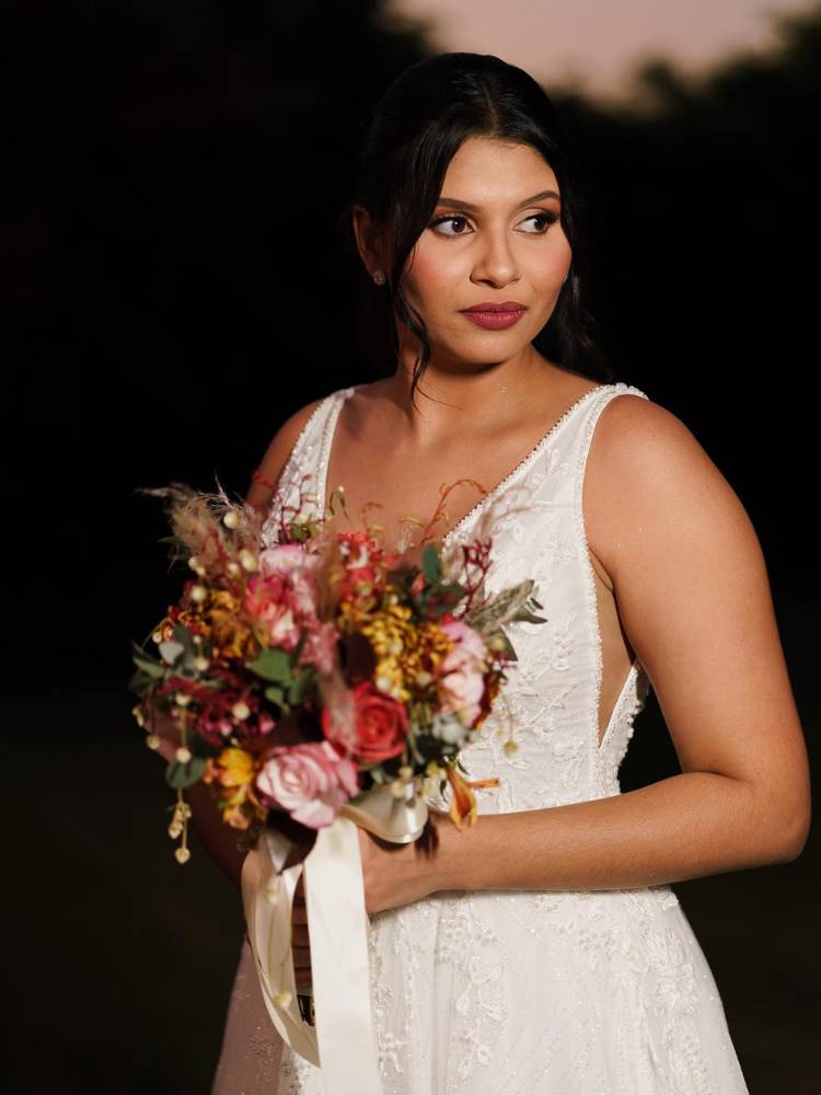 Noiva com maquiagem para casamento à noite, usando vestido branco com bordado, buquê de flores colorido em fundo preto