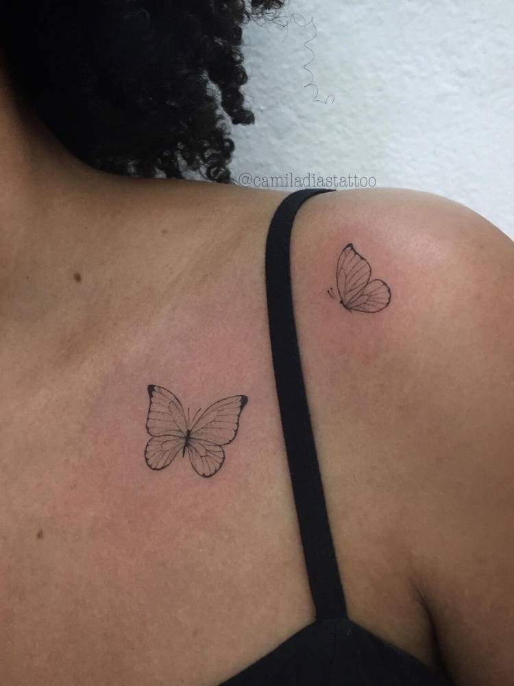 Duas pequenas borboletas em traço fino no ombro de uma mulher de pele morena. 