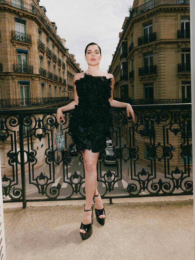 Influenciadora Gkay de pele clara com um vestido preto floral curto e sem alças. Também usa salto alto preto e a paisagem de fundo são prédios parisienses. 