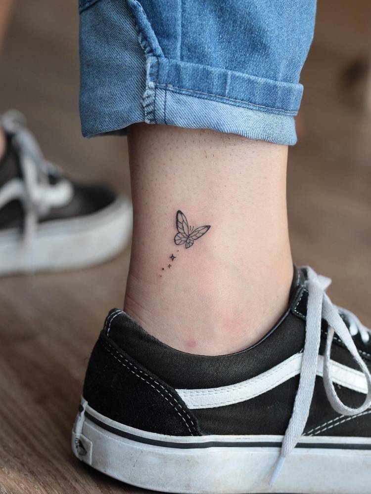 Tatuagem pequena de uma borboleta em traços finos no tornozelo de uma mulher de pele clara usando calça jeans e tênis preto.