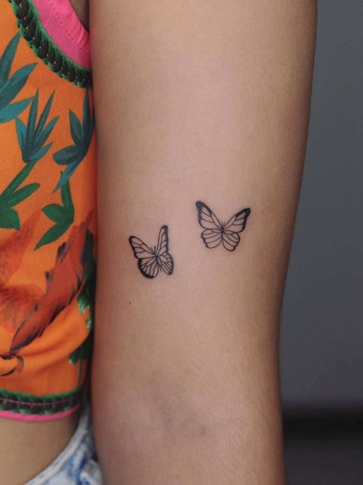 Tatuagem de duas borboletas pequenas, delicadas com traço fino no interior do braço de uma mulher de pele clara e vestindo uma blusa laranja e verde.