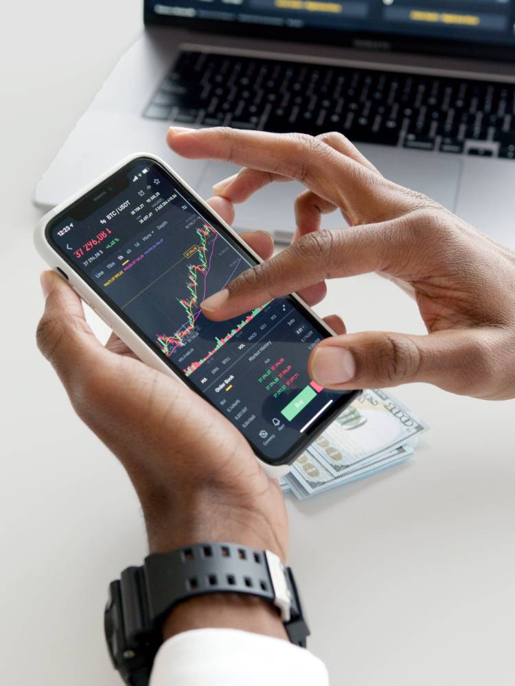 Mãos de um homem de pele negra segurando um celular. Na tela do aparelho, aparecem gráficos de investimento.