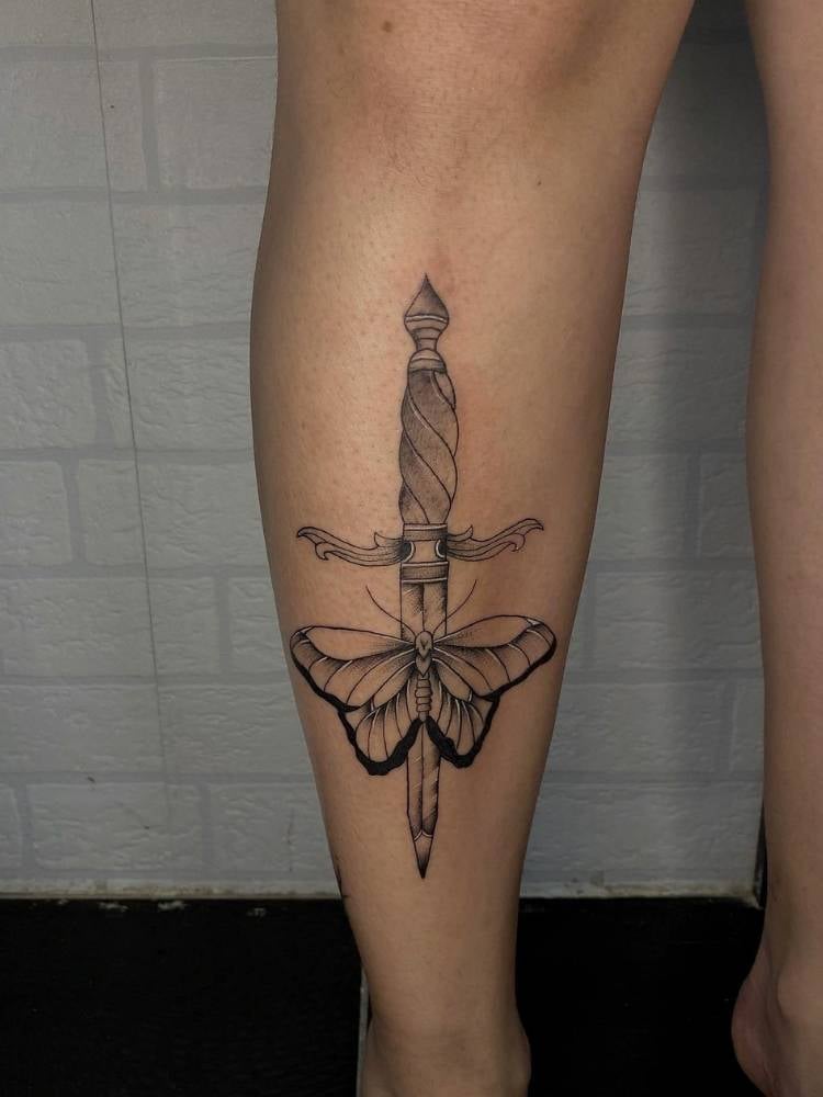 Tatuagem de borboleta com traços finos e grossos em uma perna de uma mulher de pele clara. A tattoo também apresenta uma adaga no meio da borboleta. 