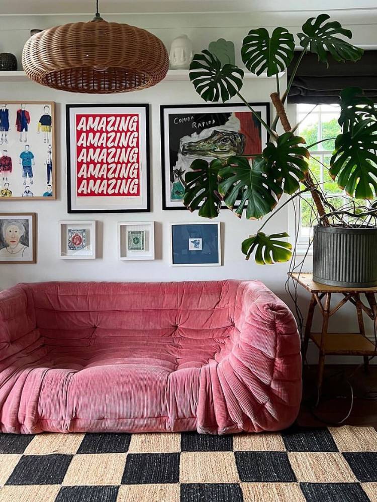 Sala com sofá rosa, vários quadros coloridos distribuídos pela parede ao fundo e grande vaso cinza com costela de adão no canto direito. 