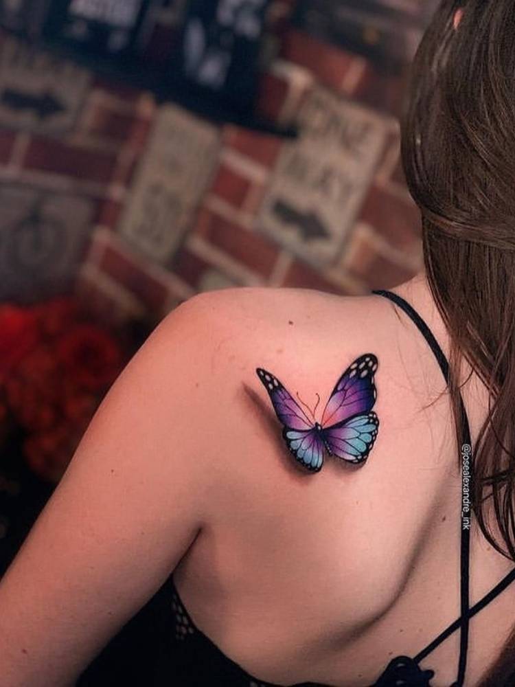 Tatuagem de borboleta roxa e azul com sombreamento na parte de trás do ombro de uma mulher de pele clara. 