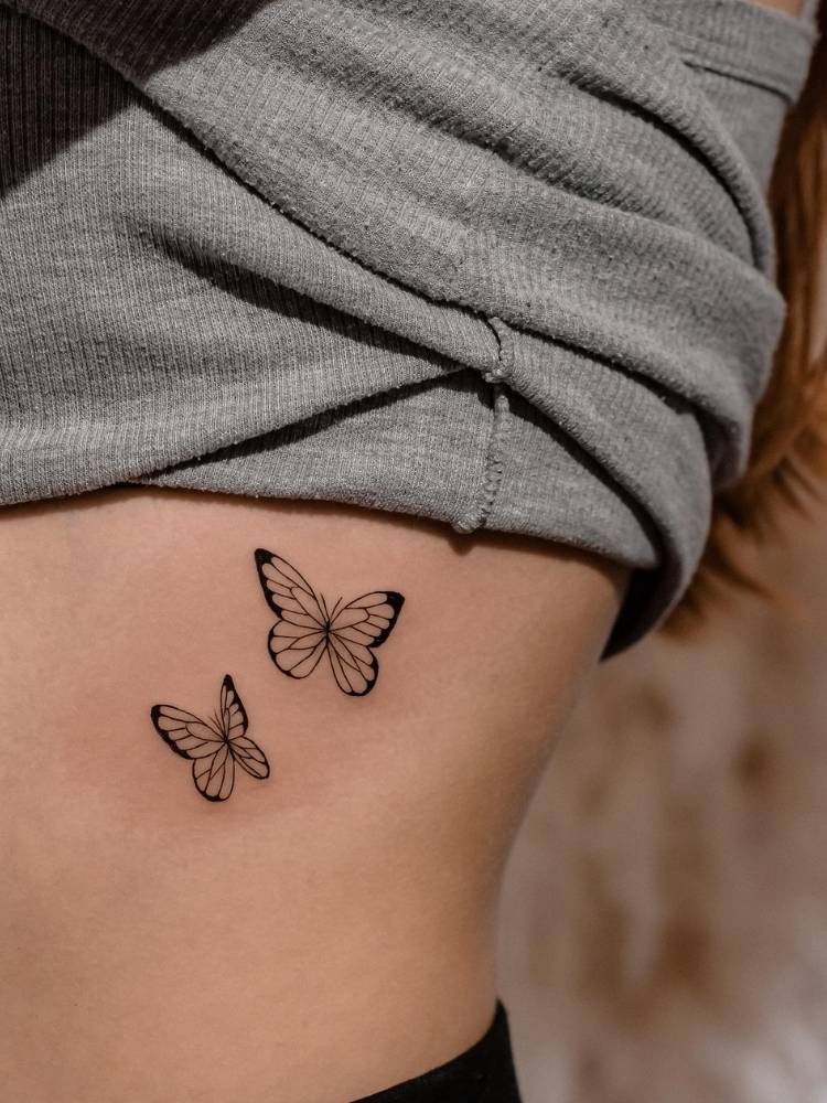 Tatuagem de duas borboletas em traço fino com um leve traço grosso na costela de uma mulher de pele clara usando uma blusa cinza. 