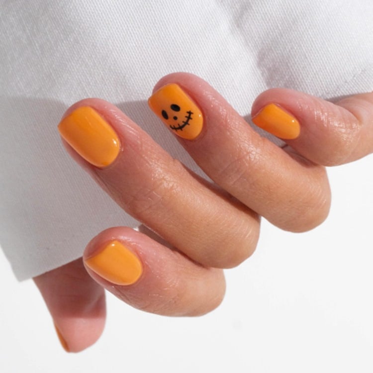 Mão com unhas curtas quadradas pintadas de laranja com carinha de abóbora de preto no dedo anelar 