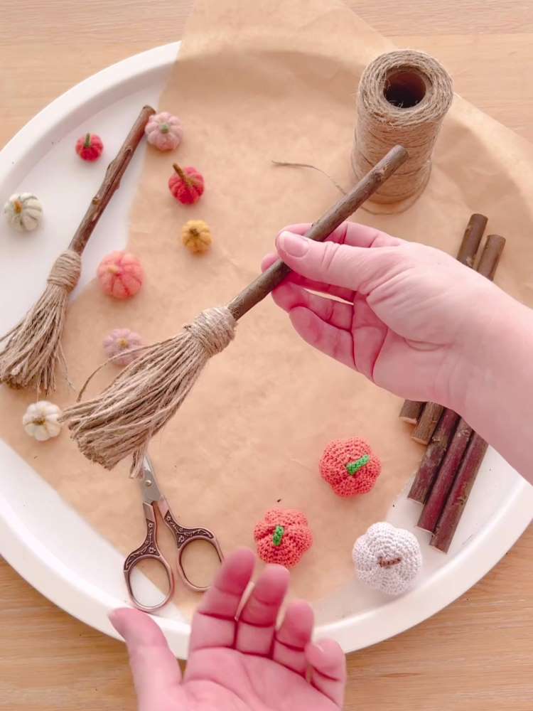 Mão segurando mini vassouras de bruxa para decorar a casa no Halloween. Fundo de abóboras de crochê, tesouras e materiais usados para o DIY