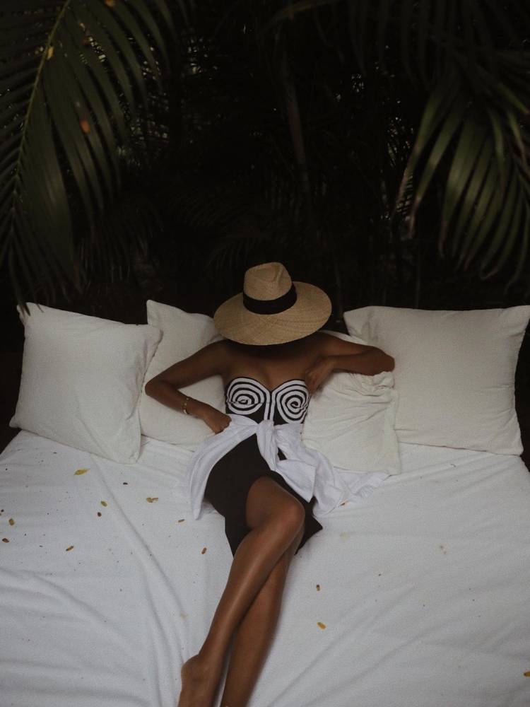 Mulher deitada sobre uma cama com lençóis brancos, usando vestido sem alça branco e preto curto e blusa branca amarrada na cintura. Ela também usa um grande chapéu. 