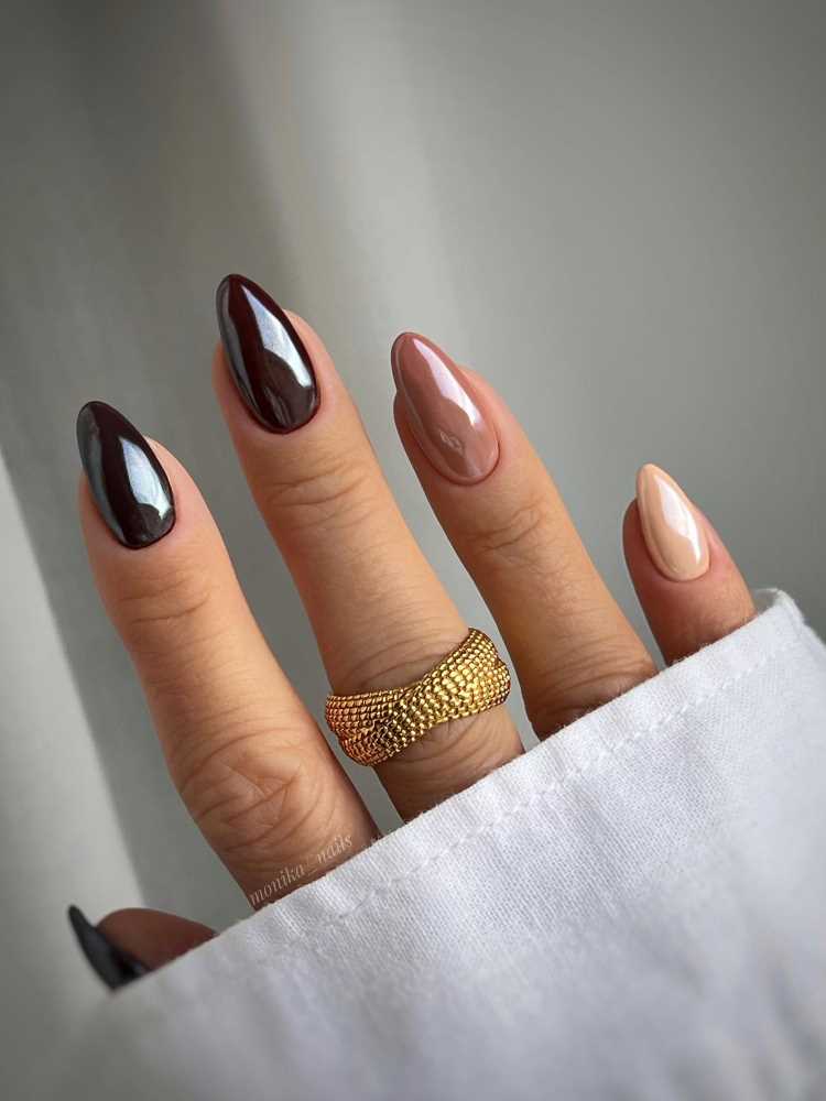 A imagem mostra a mão de uma pessoa com unhas longas e pontudas. As unhas são pintadas em tons de marrom e nude. A mão também está usando um anel de ouro no dedo médio. O fundo é uma parede cinza.
