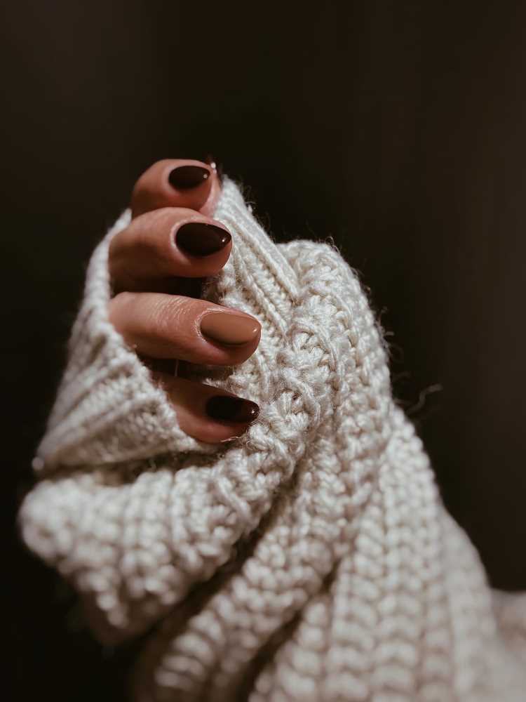A imagem mostra uma mão com unhas pintadas de escuro segurando uma blusa branca de tricô. 