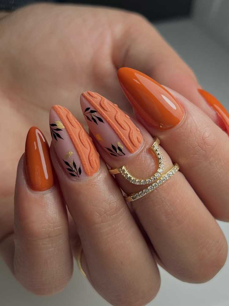 A imagem abaixo mostra uma mão com unhas longas e laranjas. As unhas têm desenhos em preto e dourado. A pessoa também está usando um anel dourado com pequenos diamantes.