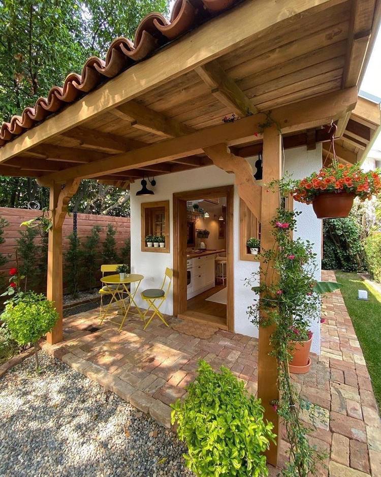 Imagem de fachada de casa simples, com alpendre, que mostra uma casa branca com a porta aberta mostrando o interior. Do lado de fora, há plantas e uma mesa amarela com 2 cadeiras