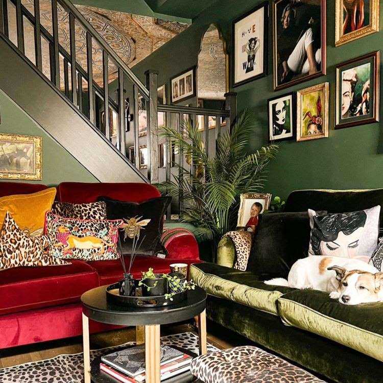 Sala de estar com paredes em verde cheias de quadro com moldura dourada, sofá vermelho de veludo, sofá verde de veludo, tapete com estampa de onça, almofadas de diversas estampas e cores. 