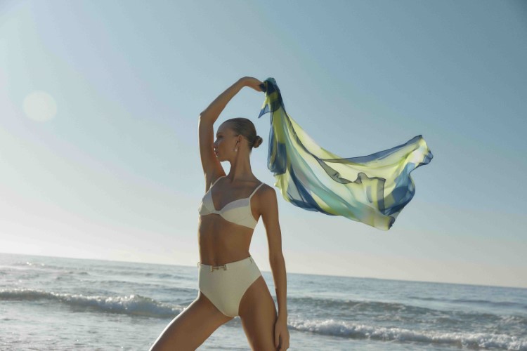 Modelo Yana Trufanova usando biquíni sutiã meia taça e parte debaixo alta, ambas na cor off white. A modelo segura um lenço verde, azul e amarelo. 