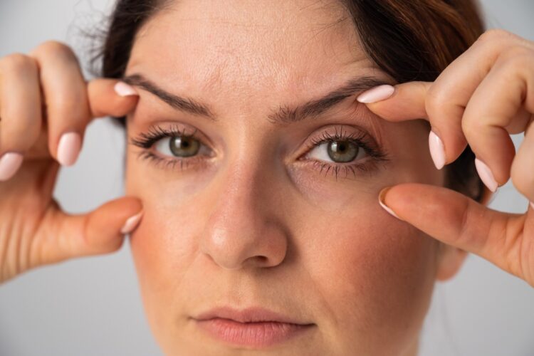 Foto de mulher de pele média e olhos verdes com os dedos indicadores e polegares esticando as pálpebras dos olhos