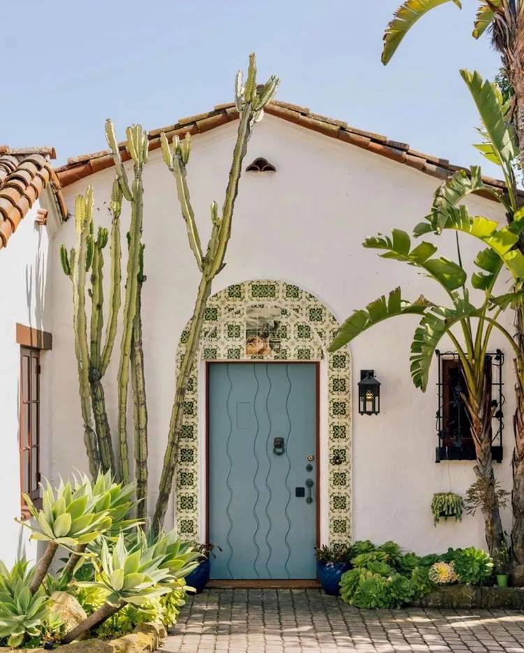 Imagem de fachada de casa simples branca, com portal decorado em azulejo, porta azul, cacto grande, bananeira e outras plantas