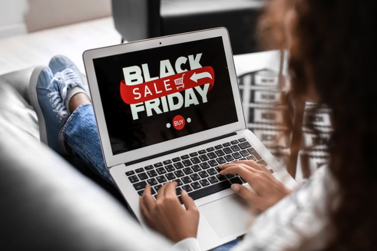 Pessoa sentada em sofá mexendo em notebook. Na tela no aparelho, está escrito em branco, vermelho e fundo preto: "Black Friday Sale Buy"