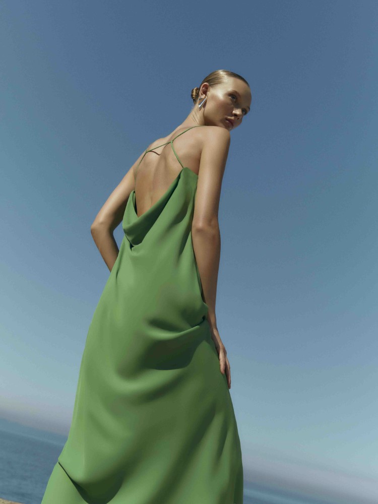 Modelo Yana Trufanova de costas usando um vestido longo verde e com tecido leve da coleção Cruise Collection 24 da Lenny Niemeyer. A parte de trás do vestido deixa as costas da modelo parcialmente aparecendo. A paisagem é o mar e um céu azul. 