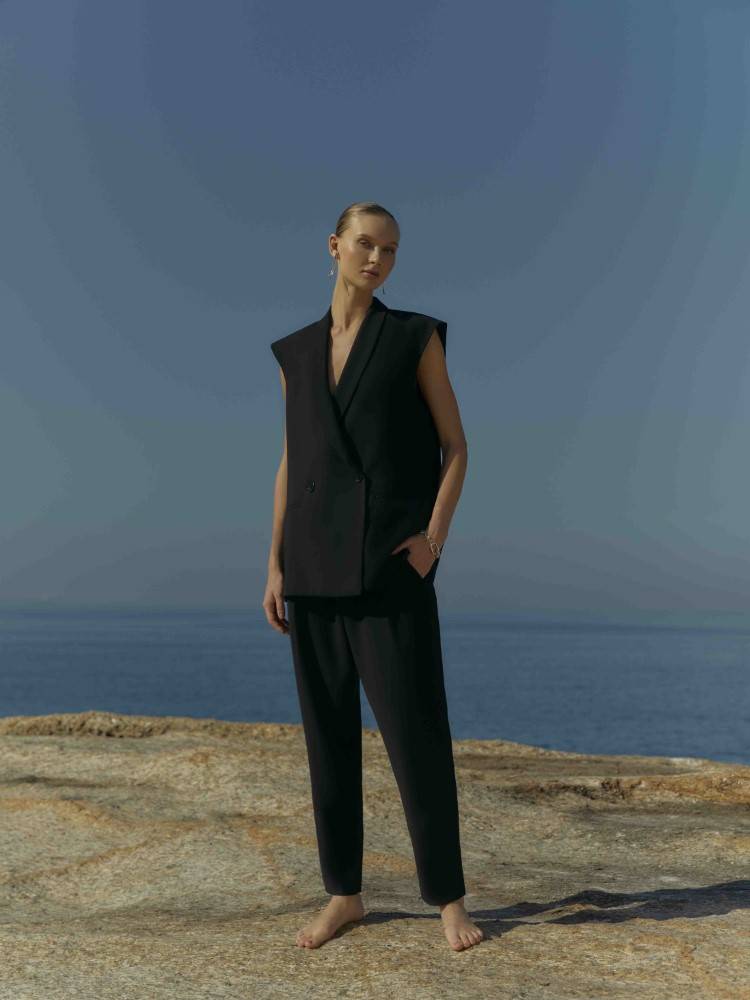 Modelo Yana Trufanova usando um conjunto de calça e blazer sem mangas na cor preta. A foto foi tirada na Pedra do Arpoador.