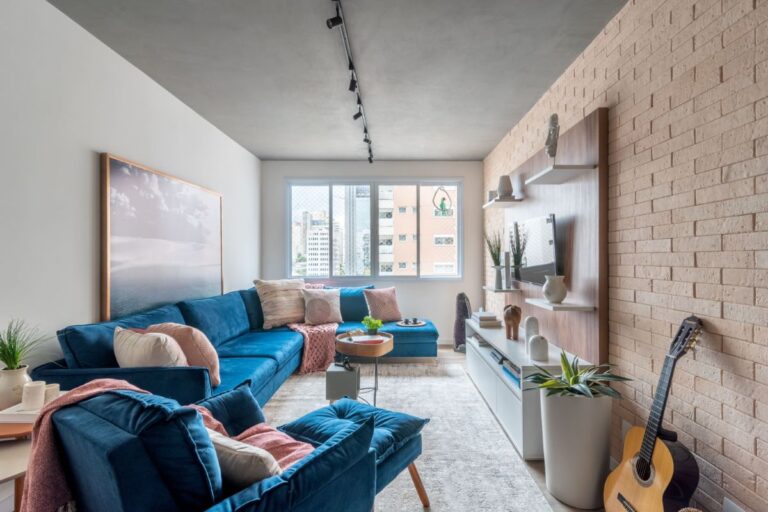 Sala de estar com sofá grande azul, poltrona também azul, almofadas rosa claro, televisão na parede, quadro atrás do sofá e ampla iluminação natural vinda de uma enorme janela.