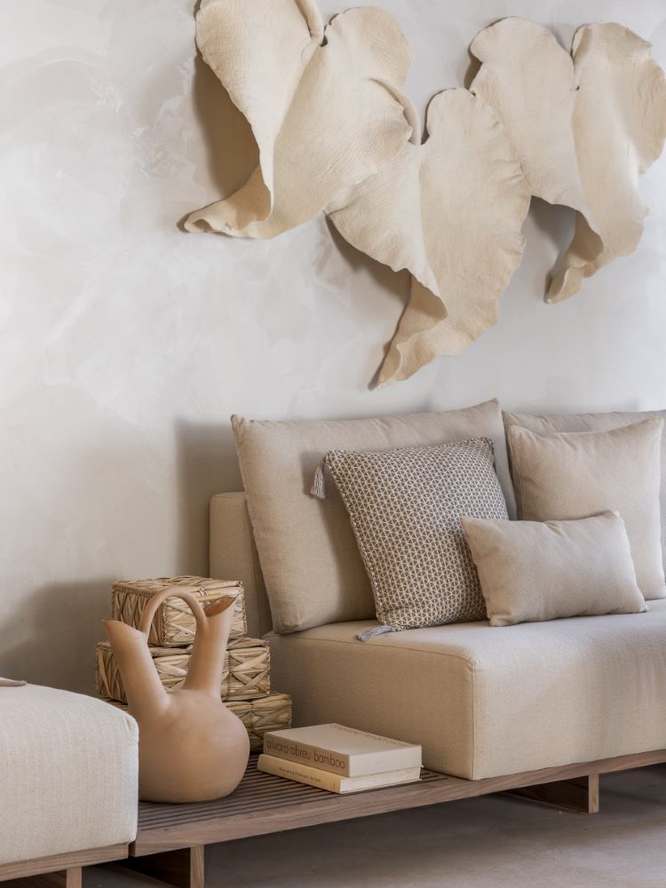 A imagem mostra parte de um sofá bege, com vaso de cerâmica ao lado e folhas artesanais na parede acima.