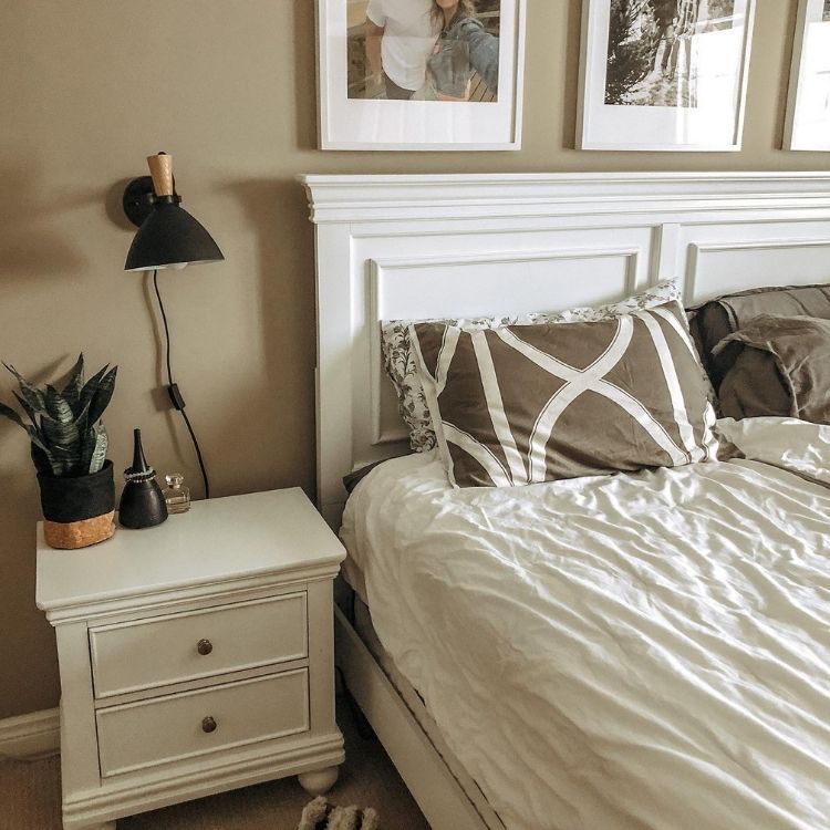 uma cama com lençóis brancos e travesseiros marrons, uma mesa de cabeceira branca com uma luminária e um vaso de planta