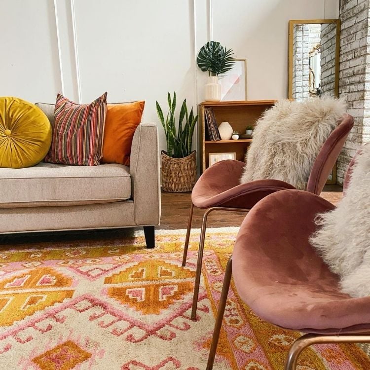 uma sala com duas poltronas rosas, um sofá rosa claro com almofadas coloridas, ao fundo dois vasos de planta. O do chão é uma espada-de-são-jorge enquanto o de cima do móvel é uma costela de Adão