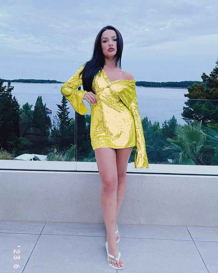 Juliette com vestido amarelo brilhoso enquanto se apoia em vidro na frente de paisagem de praia