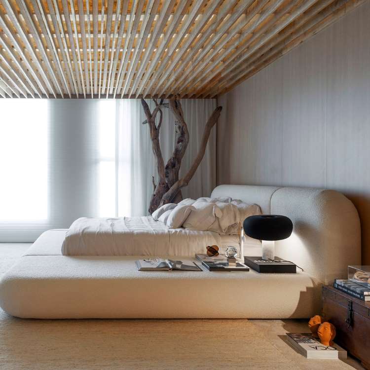 Quarto no estilo japandi, com cama baixa branca, parede em madeira, tronco de árvore ao canto e decoração minimalista. 