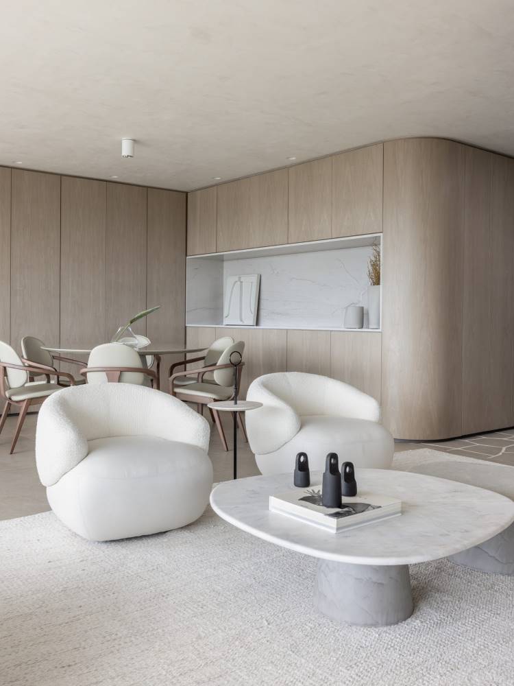 Sala de estar com poltronas brancas redondas, paredes com papel de parede que imitam madeira clara, mesas e cadeiras em tons neutros. 