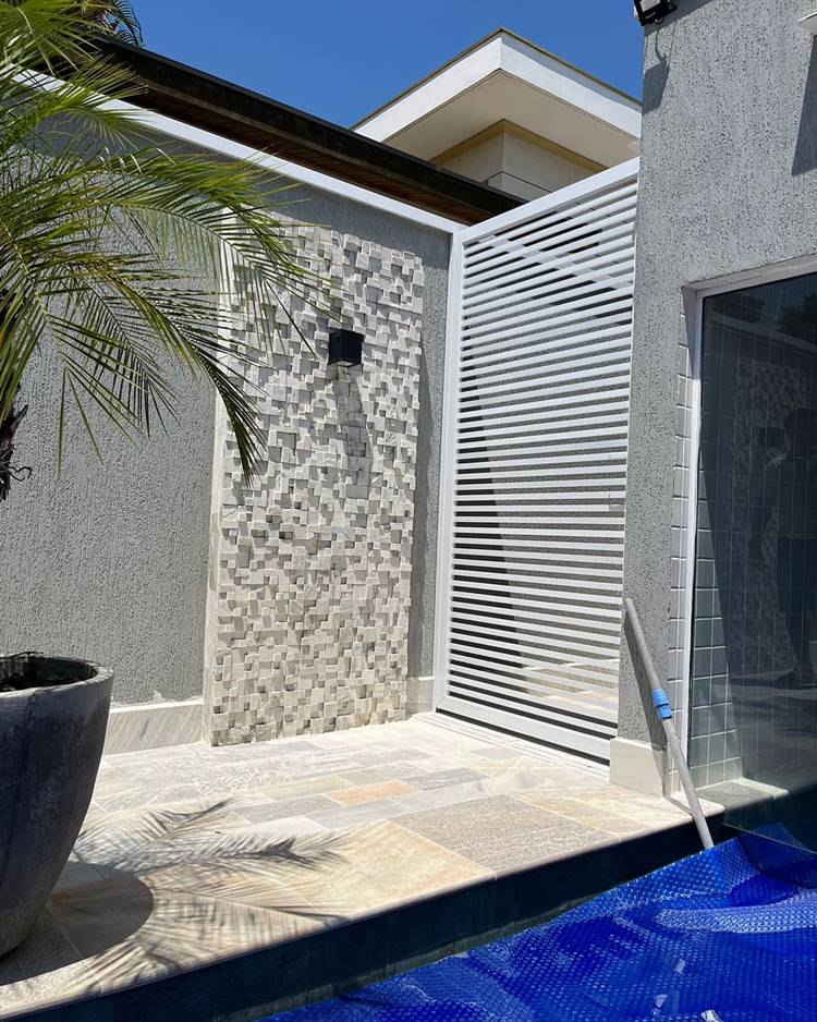 Entrada de lar com portão pequeno branco e parede detalhe 3D. De um lado uma piscina com capa azul, vaso de planta e sauna