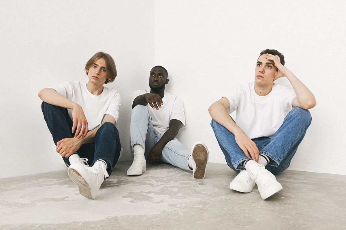 foto de três homens sentados em chão cinza usando camiseta branca, jeans masculino e tênis branco