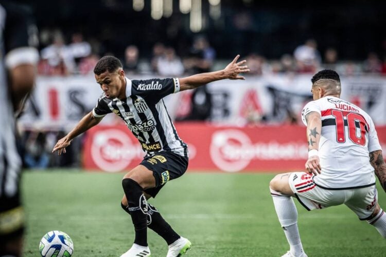 Jogador do Santos conduzindo a bola ao lado do jogador são paulino Luciano, que está de costas na foto.