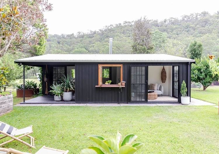 Imagem de fachada de casa simples feita em aço de cor preto. Ela está aberta, decorada com diversas plantas em vasos. Ao redor, gramado verde com cadeiras