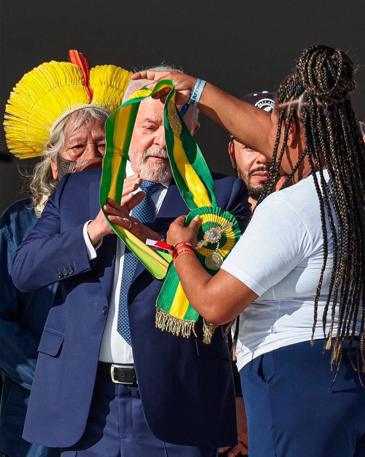 Luiz Inácio Lula da Silva recebendo a faixa de presidente da república em 2023, por mulher negra de tranças no cabelo. Ao lado do presidente, indígena e outro homem com boné.
