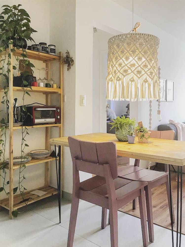 Luminária de macramê em sala de jantar com mesa e cadeira de madeira, além de estante com plantas pendentes e livros. 