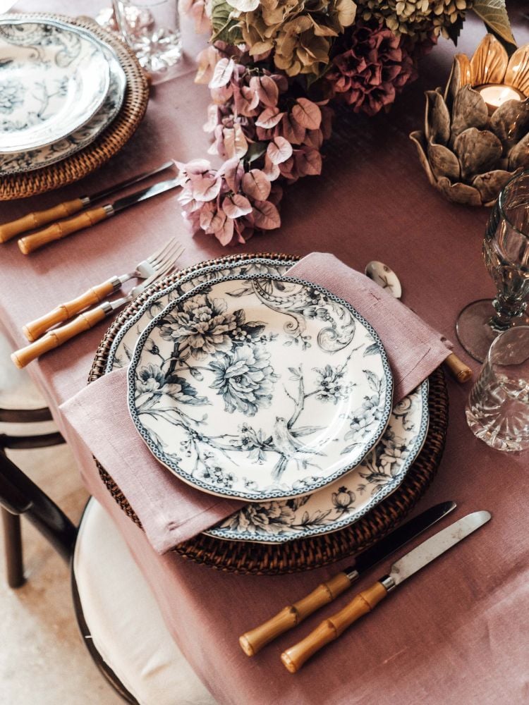 A imagem mostra uma mesa decorada com um tema floral e português. A mesa posta tem um prato branco com um desenho floral preto, talheres com cabo de bambu e um guardanapo rosa. Há também flores rosas na mesa. A toalha de mesa é de cor rosa-claro. 