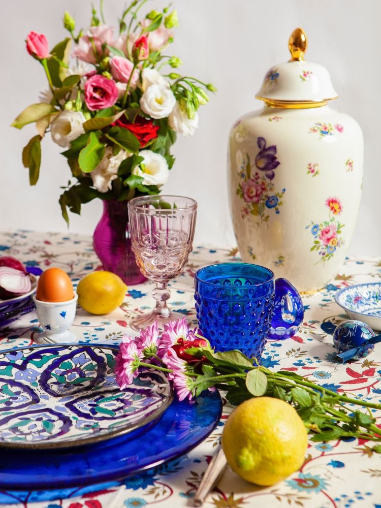 A imagem mostra uma mesa coberta por um pano branco com um padrão floral azul e vermelho. O centro da mesa é um vaso de rosas-vermelhas e brancas. Há um vaso de cerâmica branco e um desenho floral azul e rosa. Há dois copos, um é um copo de vinho transparente rosado e o outro é um copo de vidro azul. Há um prato de cerâmica azul e branco com uma flor rosa nele. Há dois limões e um ovo na mesa. O fundo é uma parede branca. A imagem transmite uma sensação de frescor e romantismo.