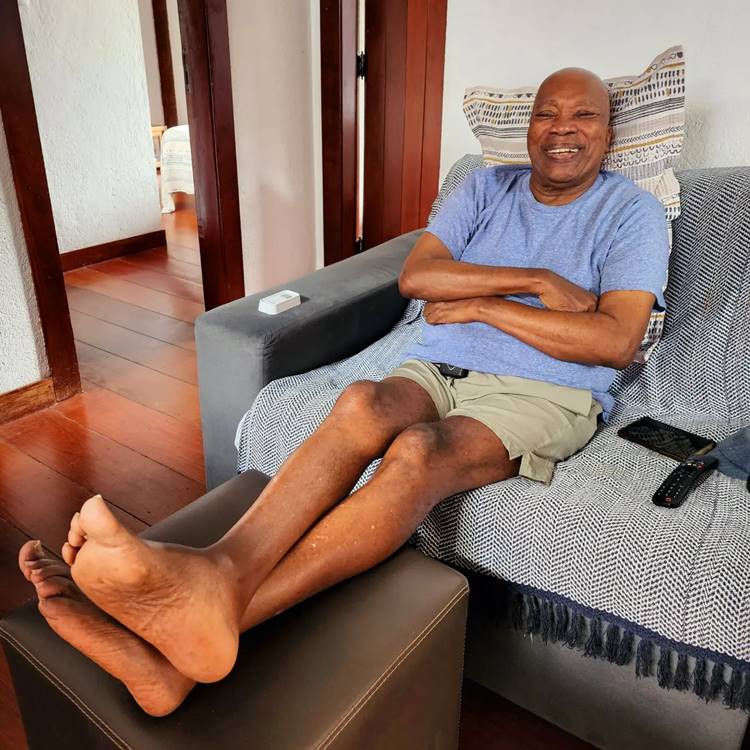 Milton Nascimento usando camiseta azul e shorts, descalço, sentado em sofá com manta. Apoiando a cabela em almofada e pés em um puf.