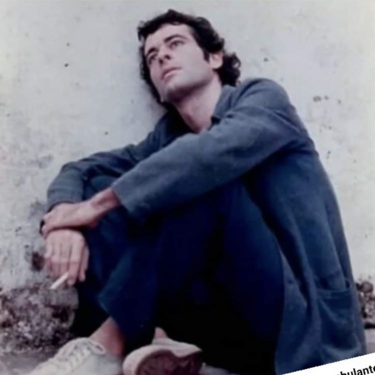 Ney antes de Secos e Molhados, em 1972, usando blazer e calça azuis, sentado apoiado em uma parede branca e com um cigarro entre os dedos. 