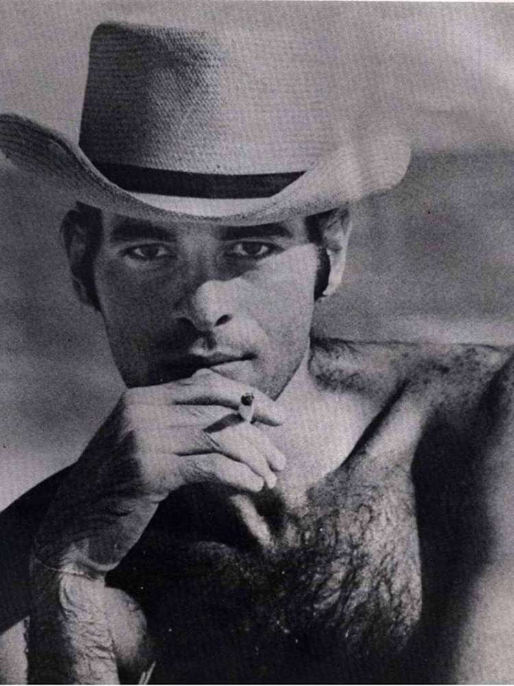 Foto em preto e branco de Ney Matogrosso em Saquarema, sem camisa, usando um chapéu e com um cigarro entre os dedos.