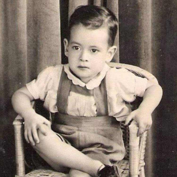 Imagem em preto e branco de Ney Matogrosso com 3 ou 4 anos, sentado em uma cadeira, de perna cruzada, usando blusa e bermuda com suspensório.