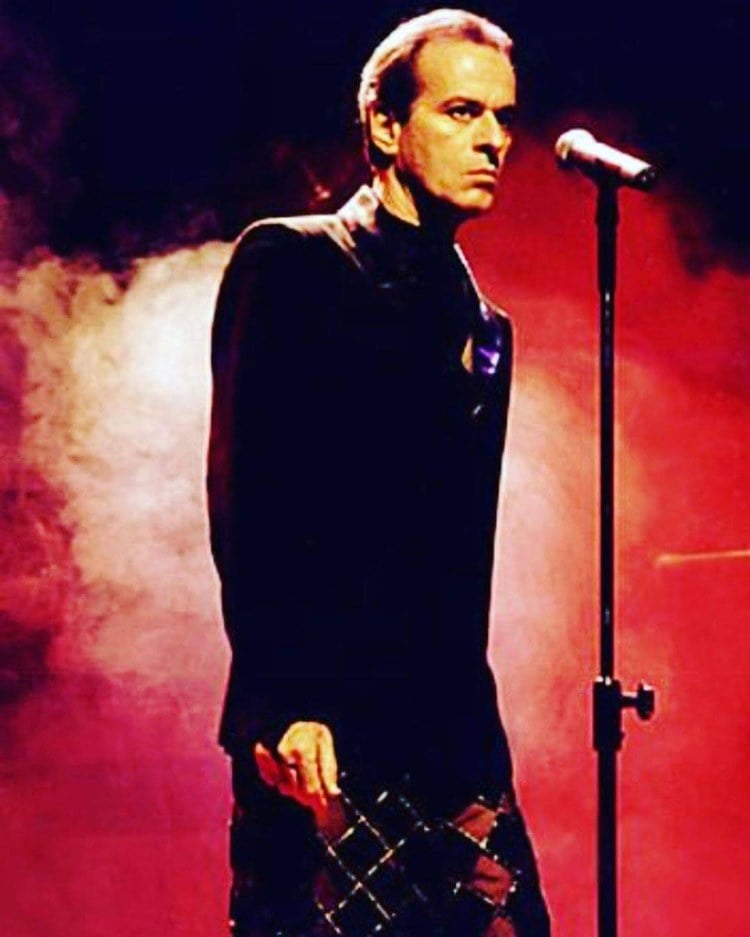 Ney Matogrosso de expressão fechada, usando um terno preto e calça da mesma cor com detalhes em vermelho e dourado. Na sua frente, há um microfone.