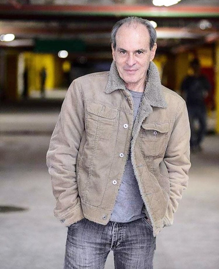 Foto de Ney Matogrosso usando jaqueta, camiseta e calça jeans em local desfocado com outras pessoas, possivelmente um estacionam,ento