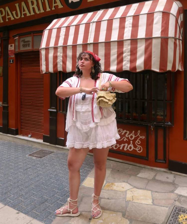 Uma mulher branca de cabelos pretos posa em frente a uma loja com um toldo listrado. Ela usa um lenço vermelho na cabeça, um vestido branco curto e papetes brancas de amarração