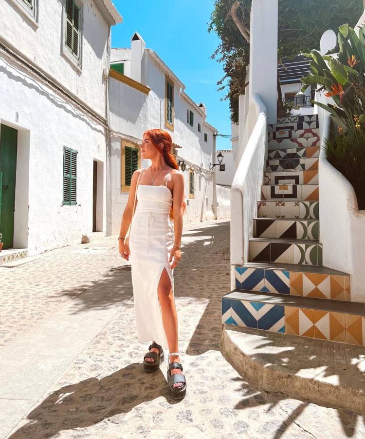 Uma mulher branca e ruiva posa em uma rua, proximo a uma escada de azulejos. Ela umas um vestido longo branco com uma fenda e uma papete preta.